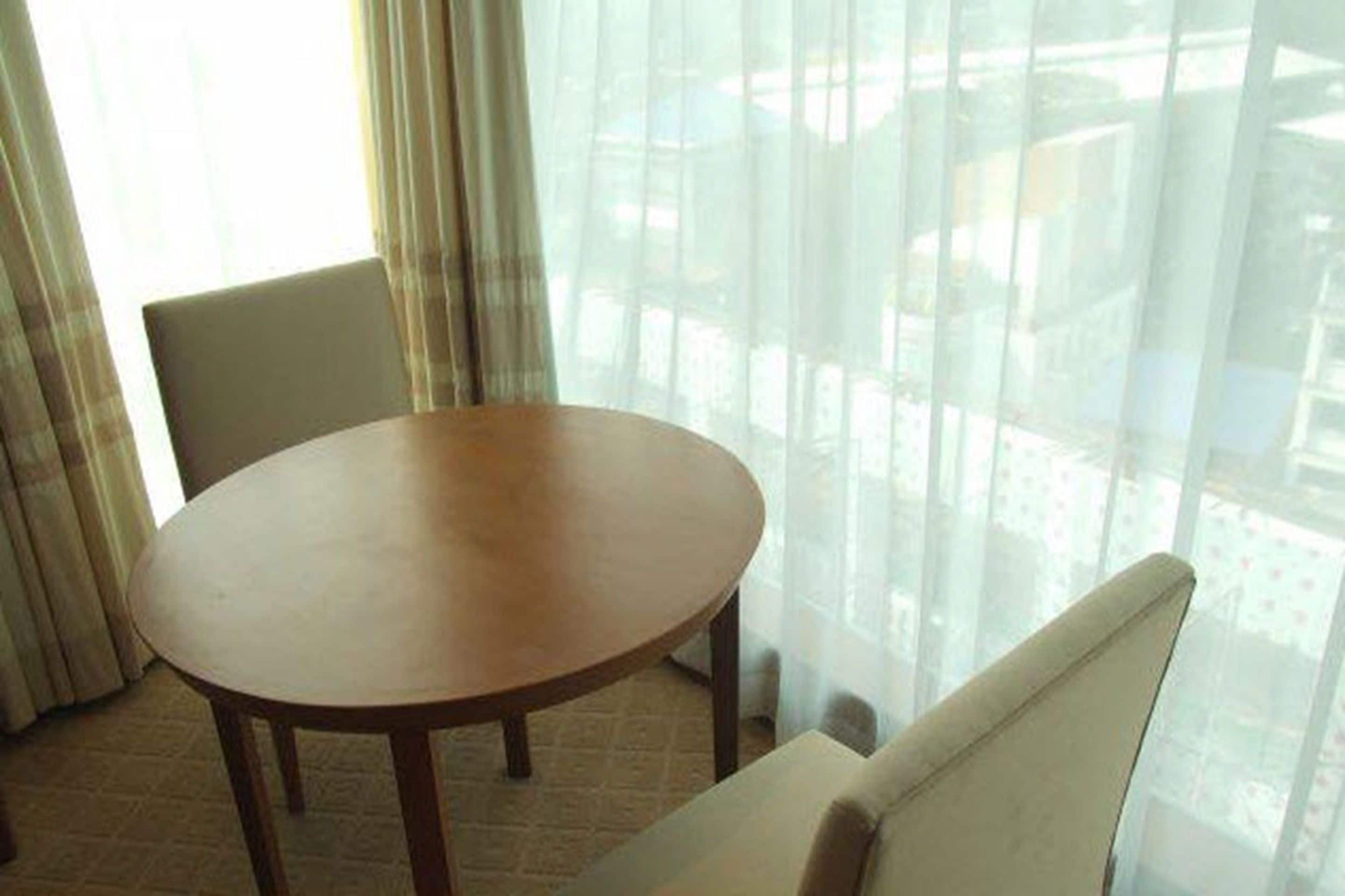 Una habitación con una mesa redonda de madera y una silla blanca, ventanas amplias con cortinas translúcidas y vistas a edificios.