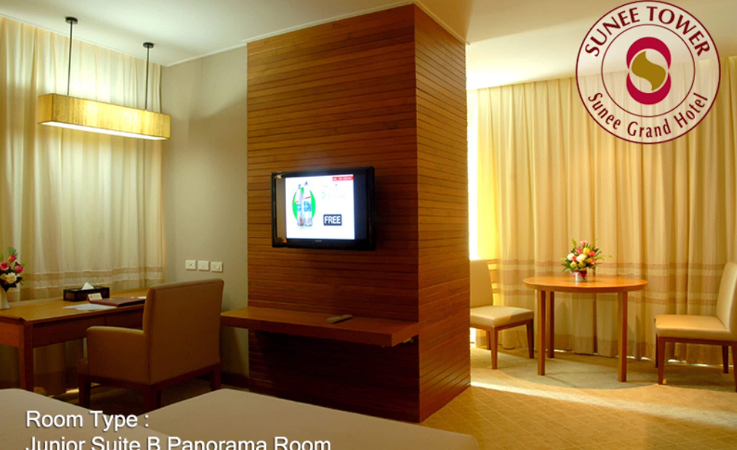 A Junior Suite B Panorama Room with a television mounted on a wooden panel, a dining area with a table and chairs, and a large window with curtains.