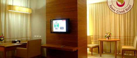 Una Junior Suite B Panorama con un televisor montado en un panel de madera, un comedor con mesa y sillas, y un gran ventanal con cortinas.