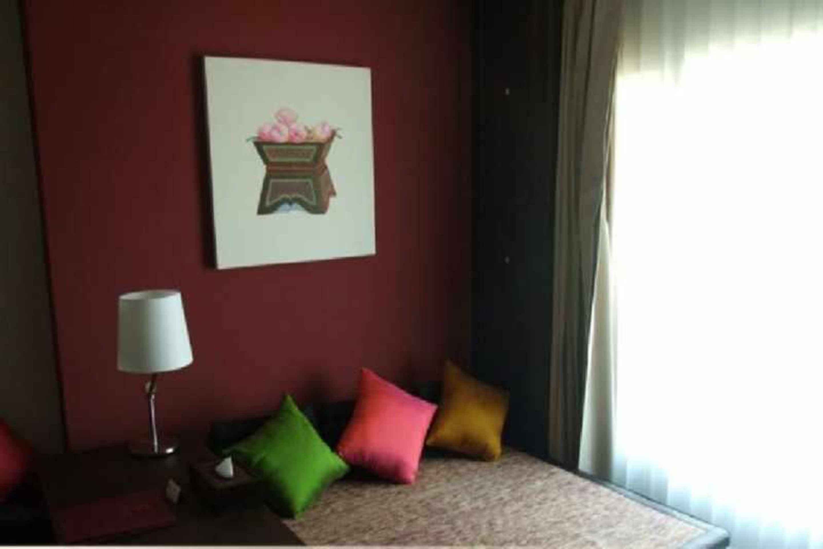 Une chambre d’hôtel avec un mur rouge, un tableau représentant un panier de fleurs, une lampe de chevet et trois coussins colorés.