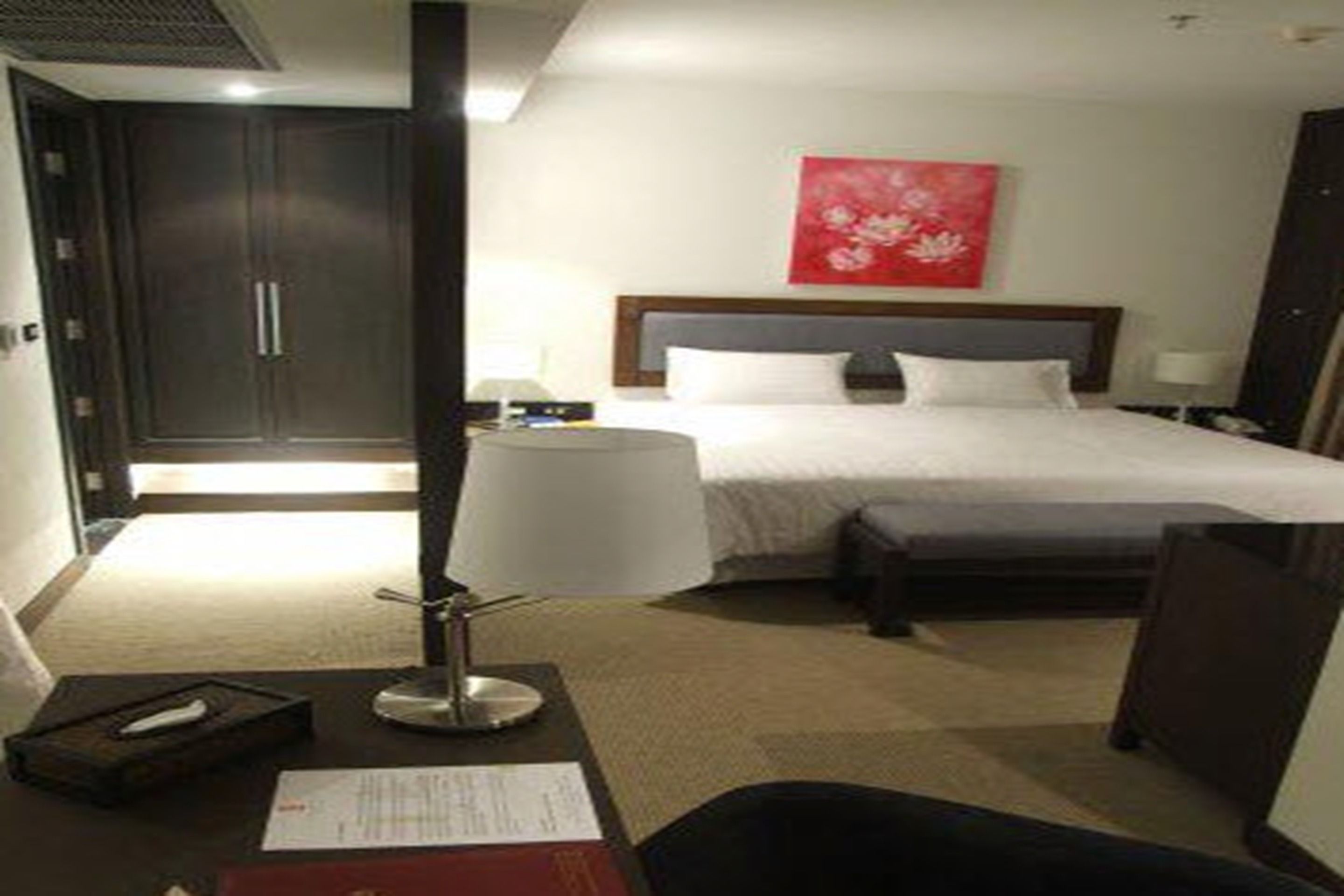 Una habitación de hotel con cama, mesitas de noche, un escritorio con lámpara, una silla y una obra de arte roja en la pared.