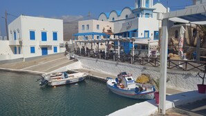 Un pueblo costero con una iglesia destacada que tiene una torre con reloj, un puerto con barcos y un edificio blanco con ventanas azules.