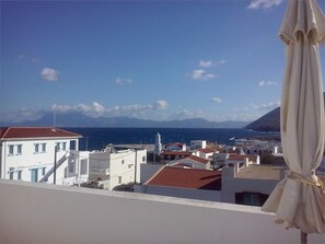 A coastal town with white buildings, a clear blue sky, and a view of the sea and mountains in the distance.
