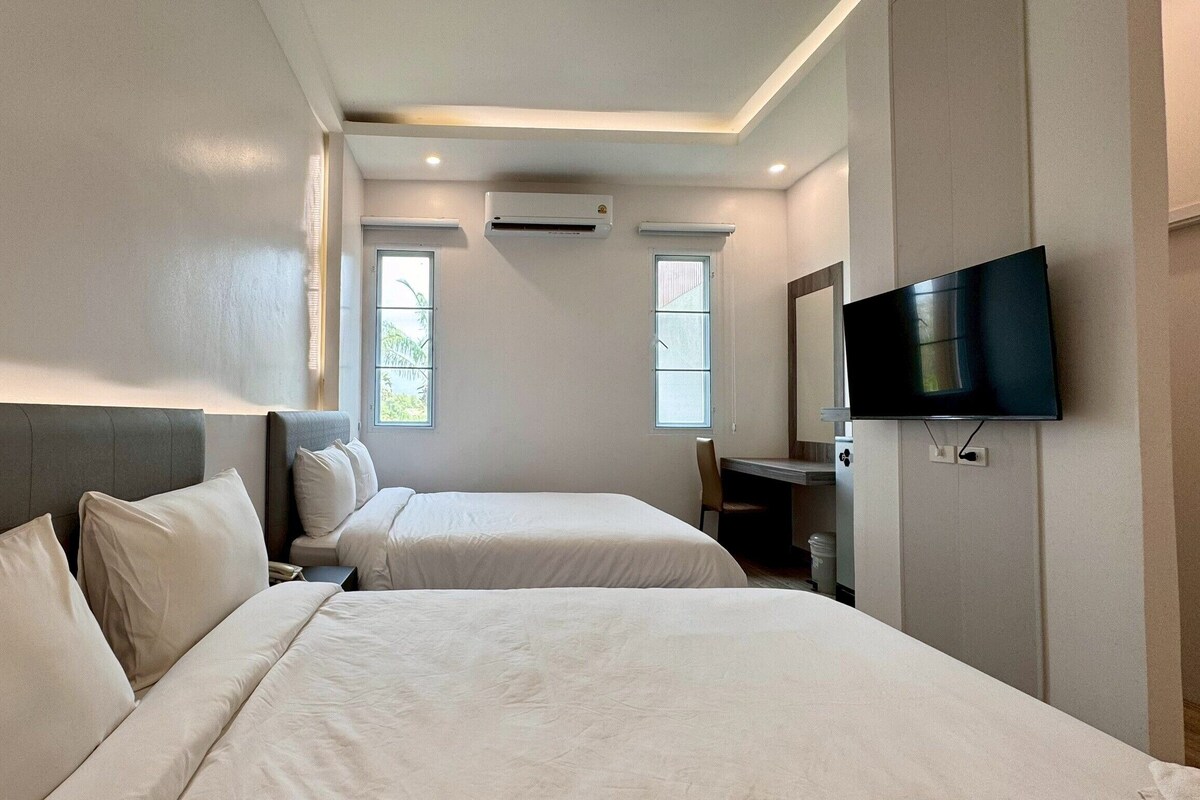 A hotel room with two beds, a desk, a chair, a TV, and two windows.