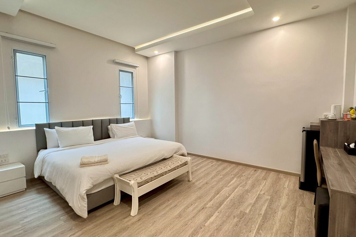 A modern bedroom with a bed, bedside tables, a bench, a TV, and two windows.