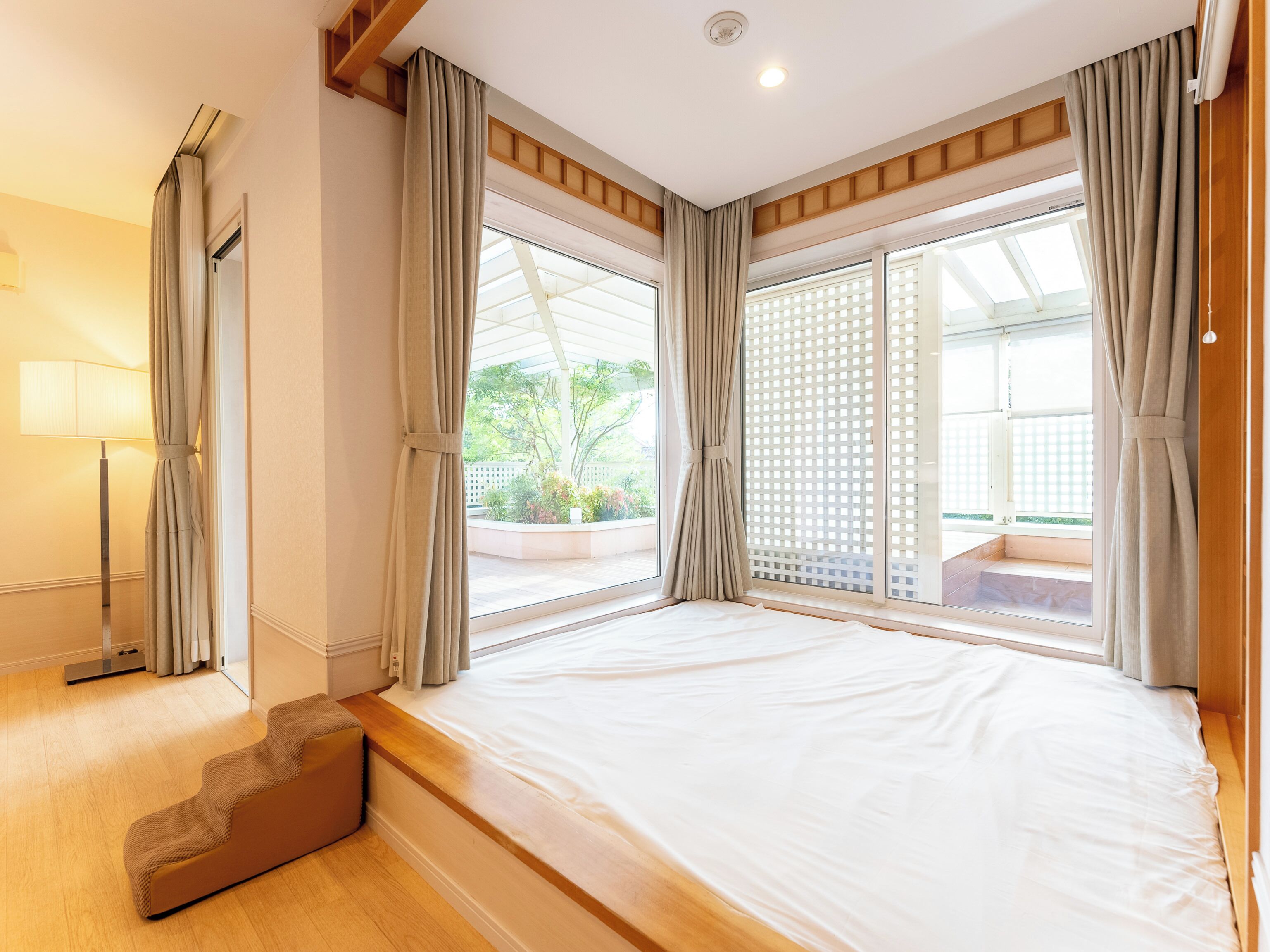 A bedroom with a large bed, a wooden bench, a floor lamp, and a view of a garden through a sliding door.