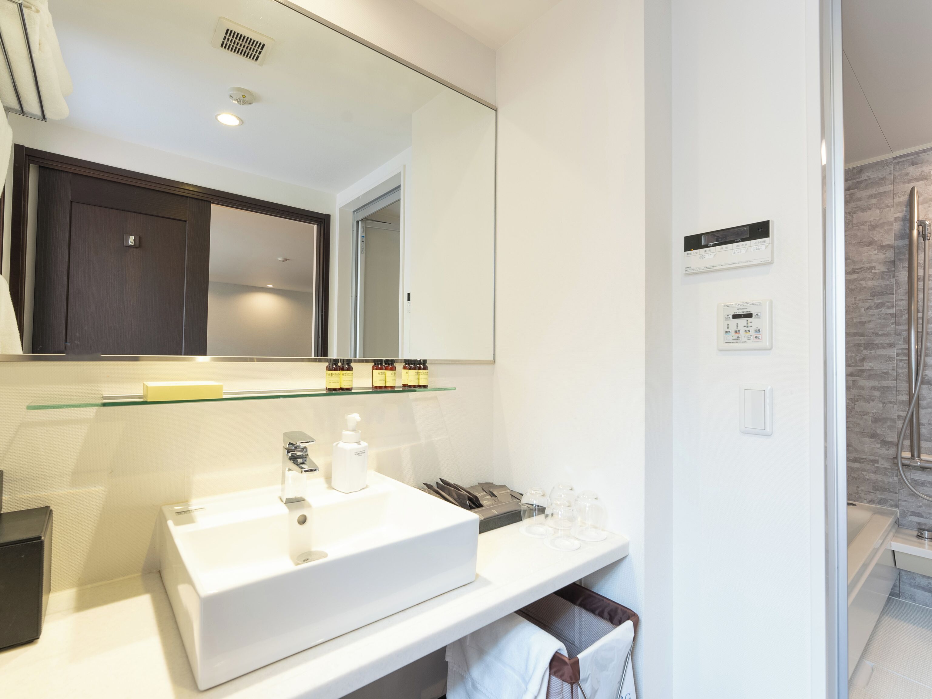 Un baño moderno con un espejo grande, lavabo blanco y estante de vidrio con artículos de aseo.
