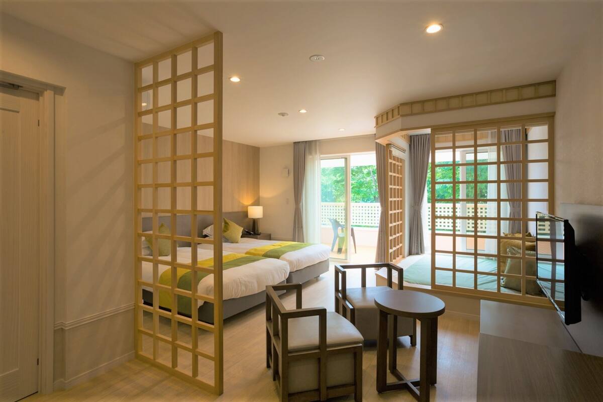 A hotel room with a bed, a desk, a chair, and a TV. There is a sliding door leading to a balcony with a view of greenery.