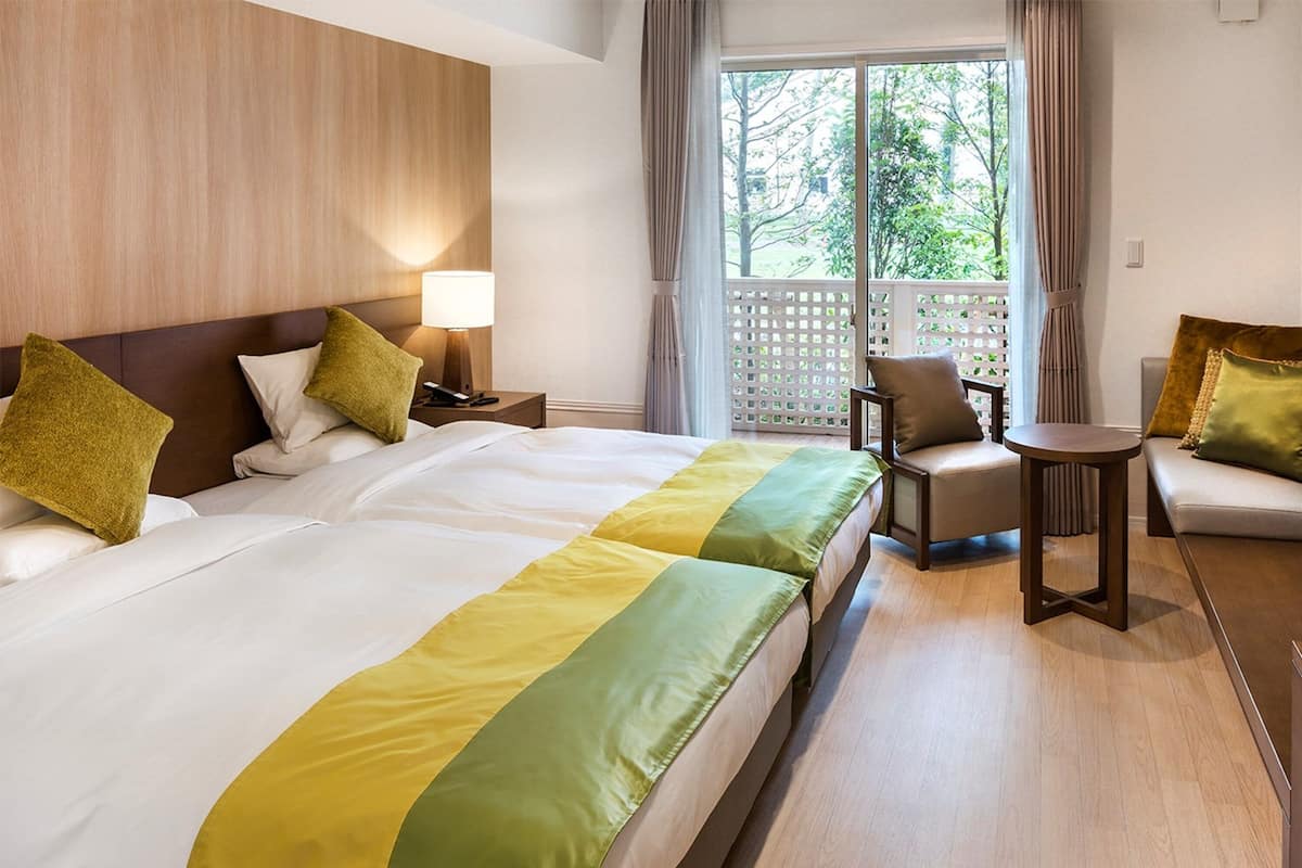A hotel room with a large bed, a chair, a small table, and a view of greenery through the window.