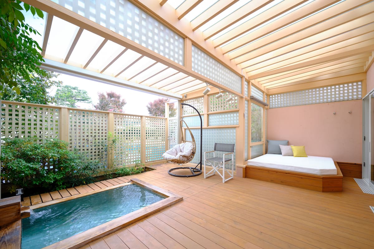 A spa-like setting with a wooden deck, a hot tub, a seating area with a swing chair, and a bed with pillows.