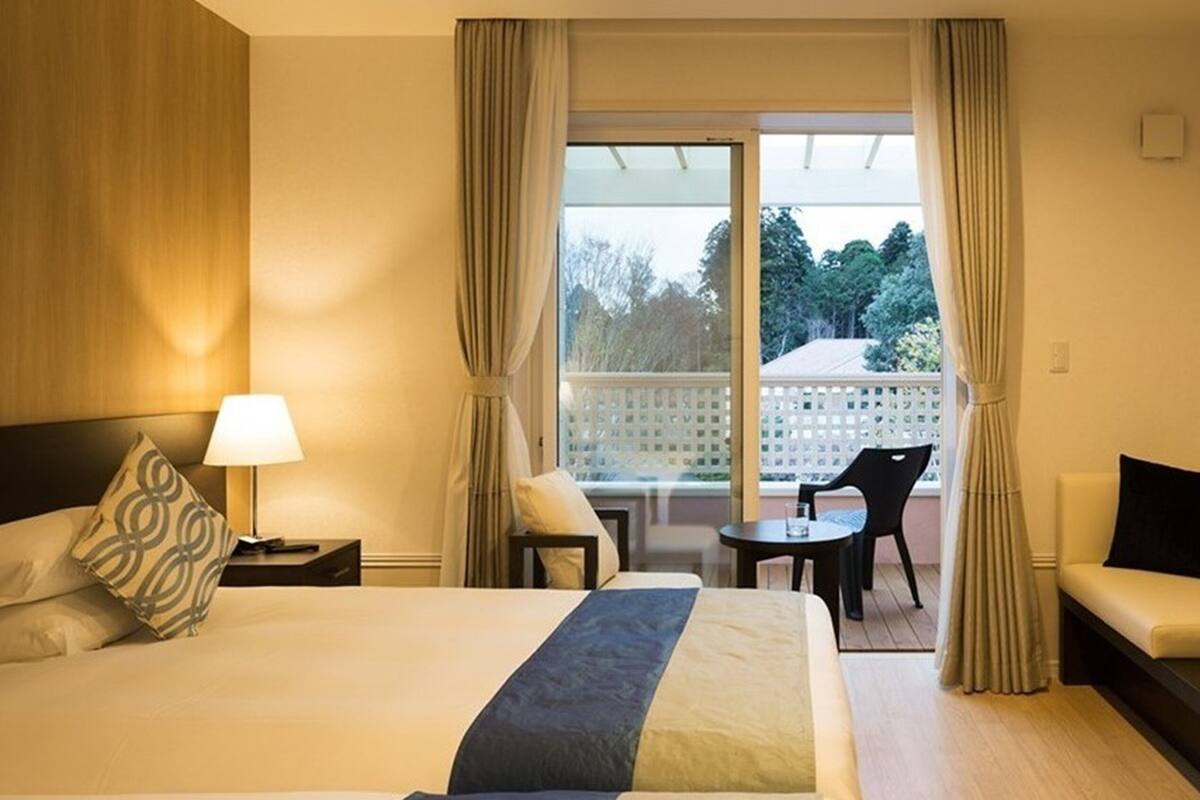 A hotel room with a large bed, a desk with a lamp, a sofa, and a balcony with a table and chairs.