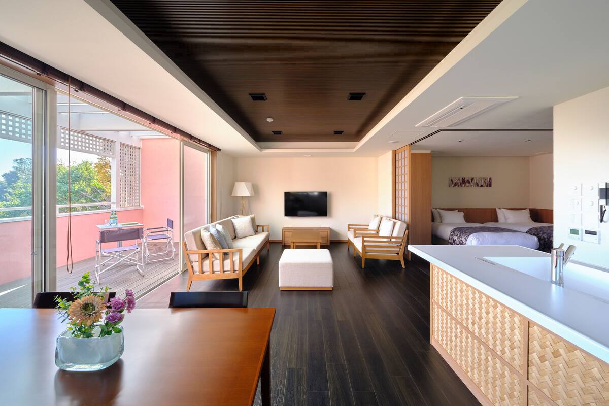 A modern hotel room with a wooden ceiling, a large window, a sofa, a desk, a bed, and a bathroom with a sink and mirror.