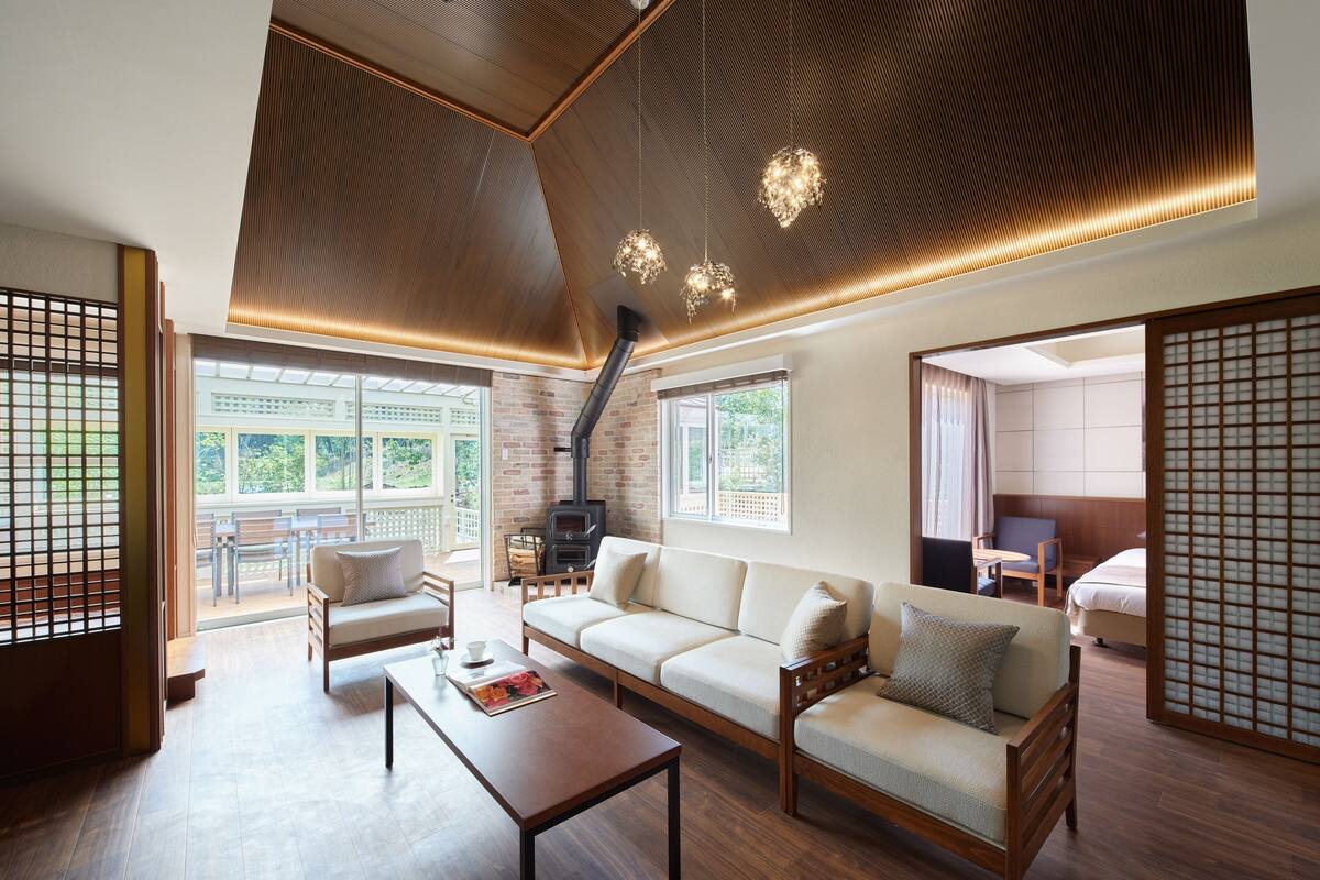 A modern living room with a sofa, coffee table, and a fireplace. There are large windows providing natural light and a view of the outdoors.