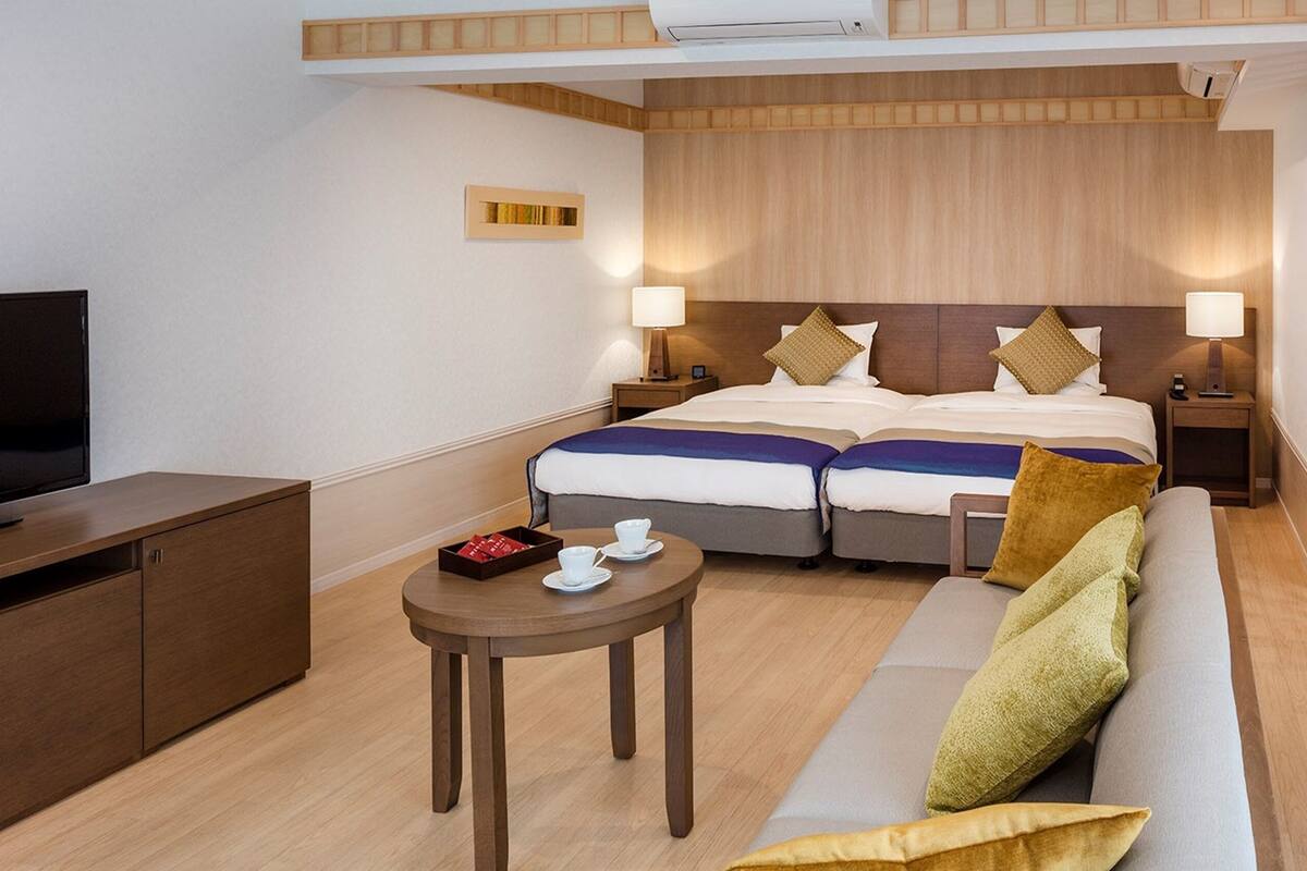 A hotel room with a bed, two bedside tables, a TV, a sofa, and a small table with cups and a box.