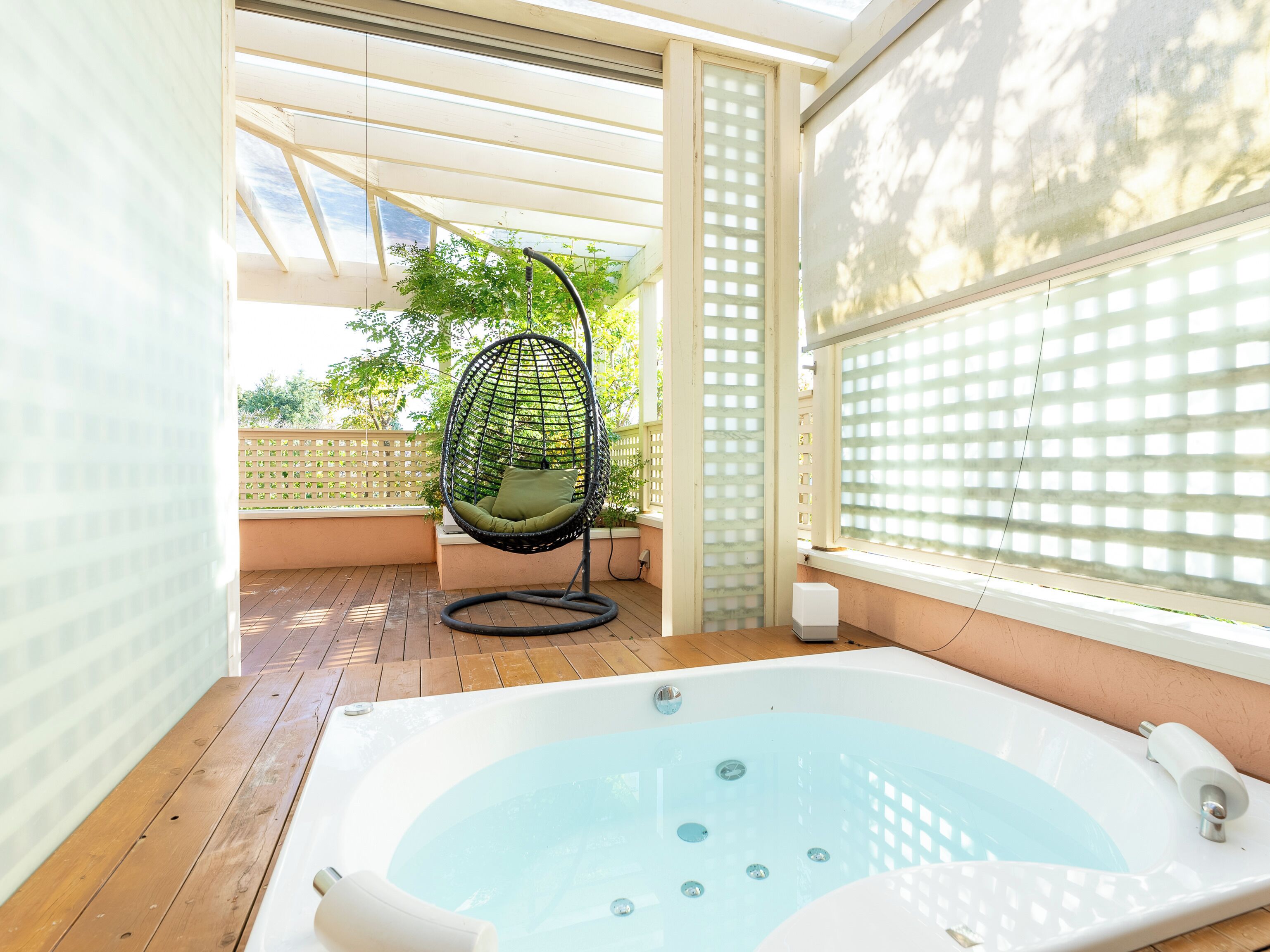 A hot tub with a wooden deck, a hanging chair, and a view of greenery through a window.