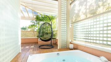 A hot tub with a wooden deck, a hanging chair, and a view of greenery through a window.