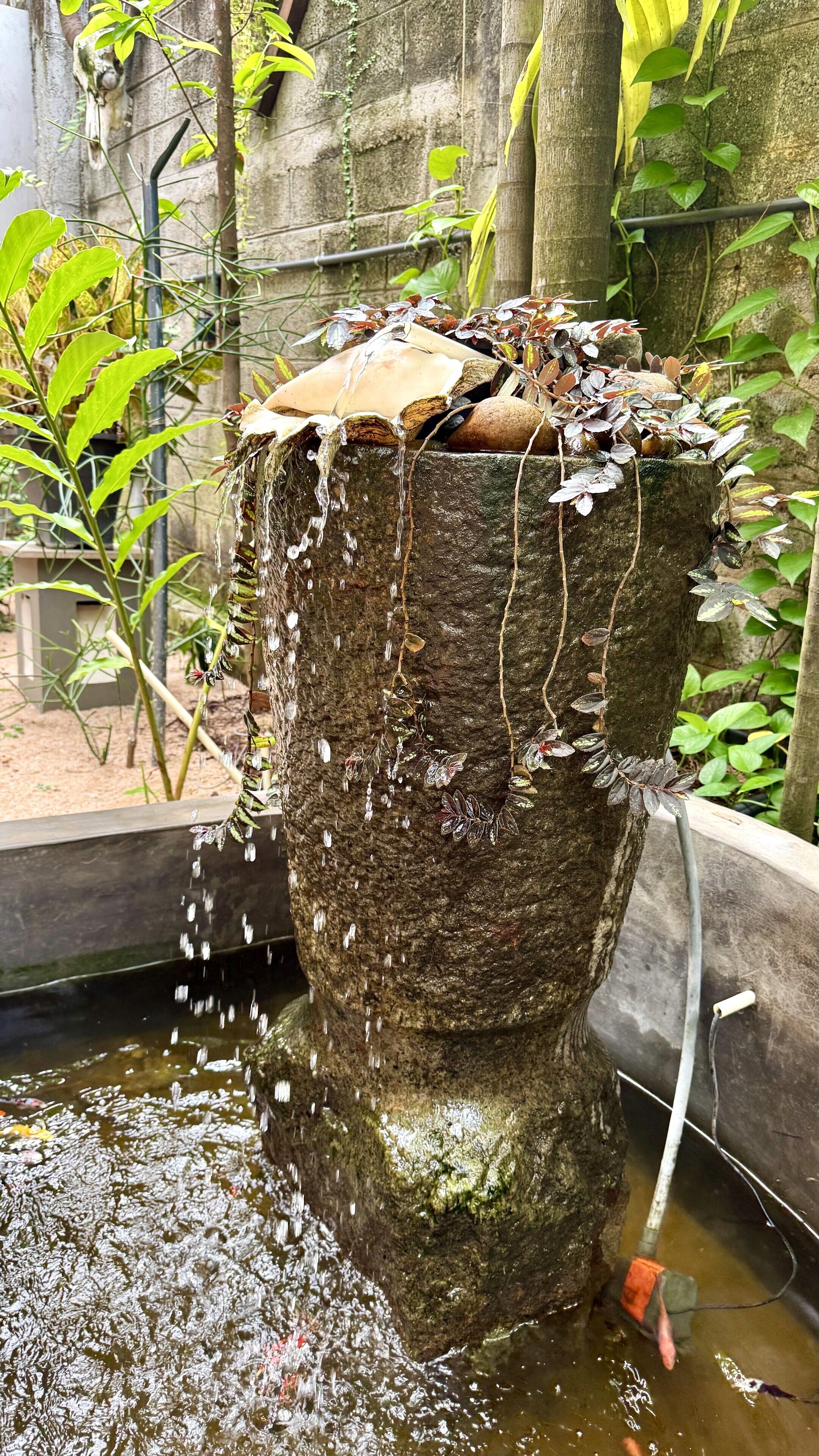 Ein Steinfountain, über den Wasser fließt, umgeben von Grün und einer Steinmauer.