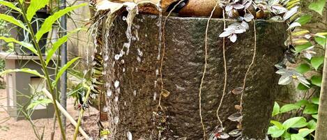A stone fountain with water flowing over it, surrounded by greenery and a stone wall.