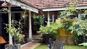 An outdoor patio area with a tiled roof, surrounded by lush greenery and a variety of plants. There are several metal tables and chairs arranged for outdoor dining.