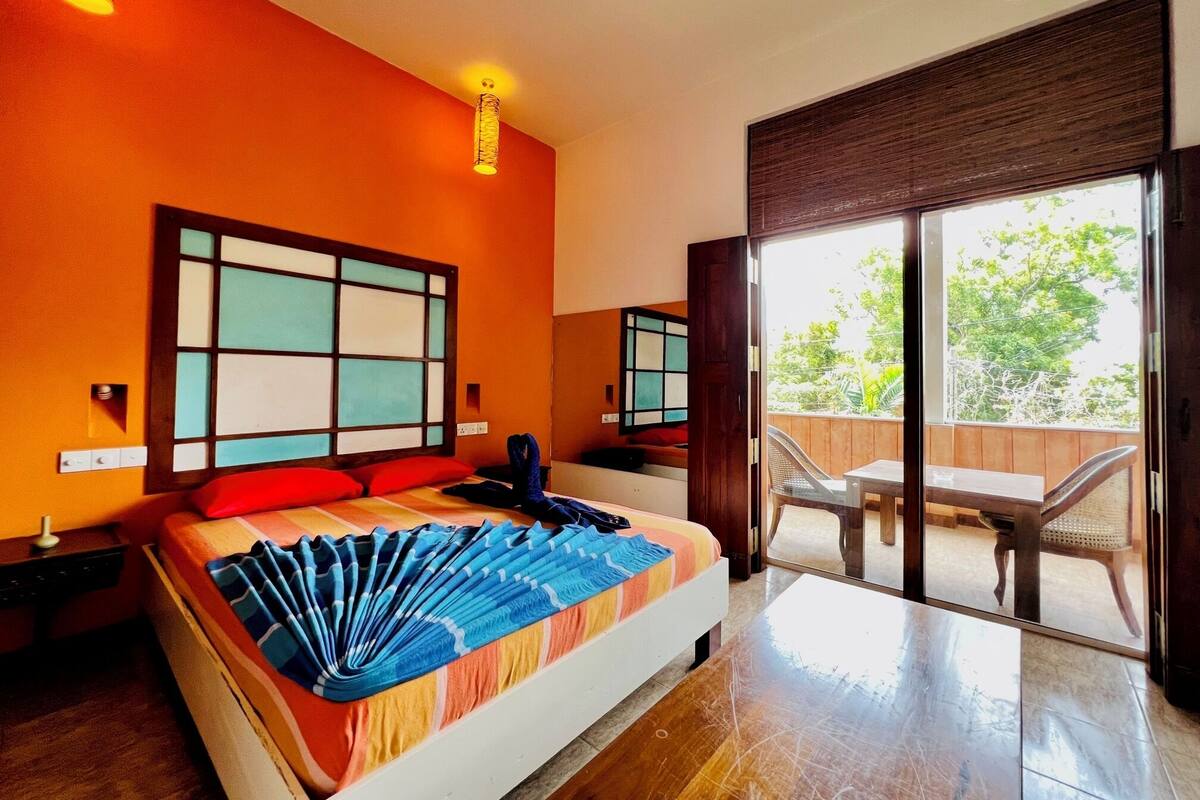 A hotel room with a large bed, a sliding glass door to a balcony, and a view of greenery outside.