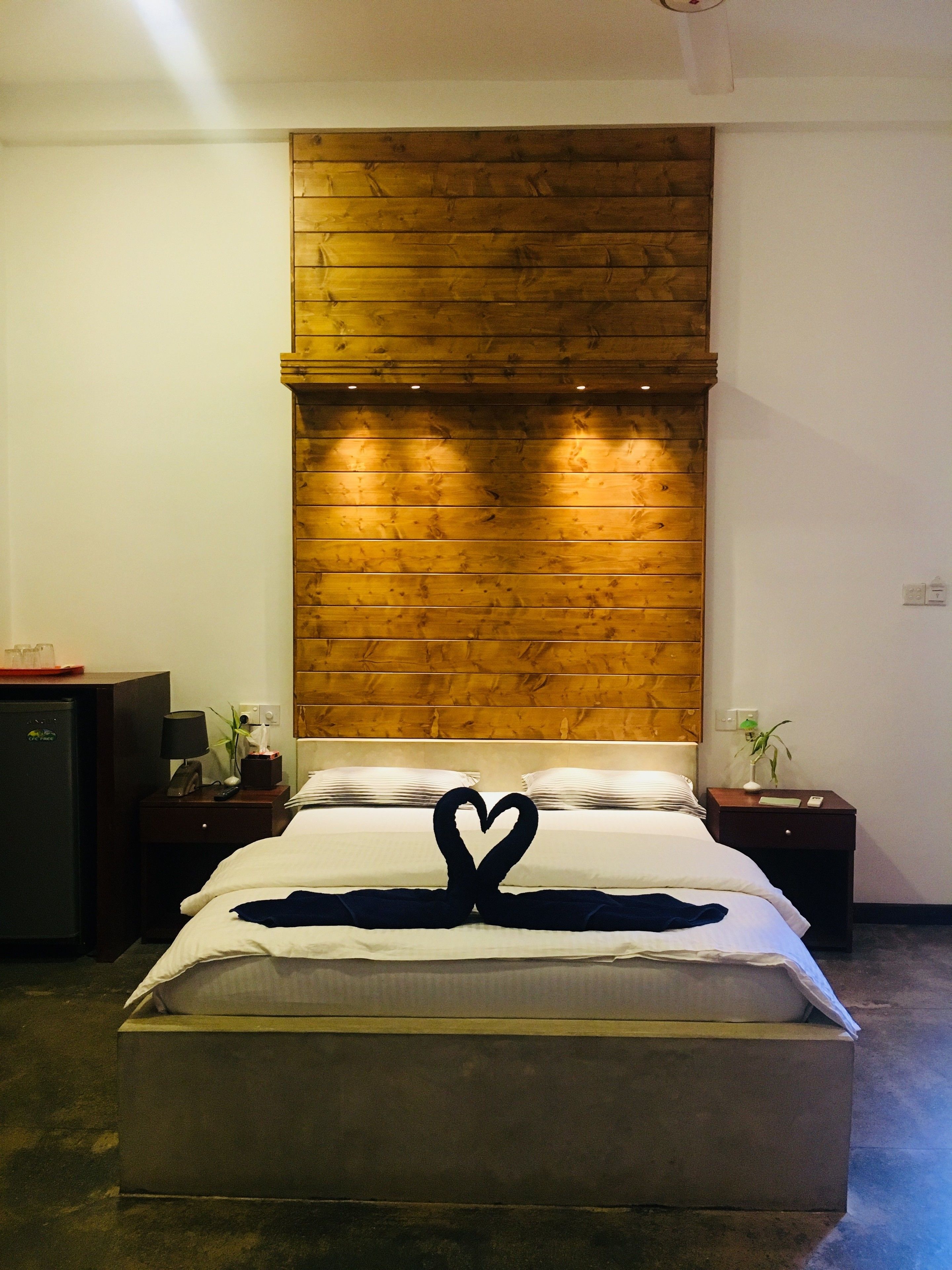 A hotel room with a wooden headboard, a bed with white linens, a heart-shaped decoration on the bed, a nightstand with a lamp, and a small plant on the nightstand.