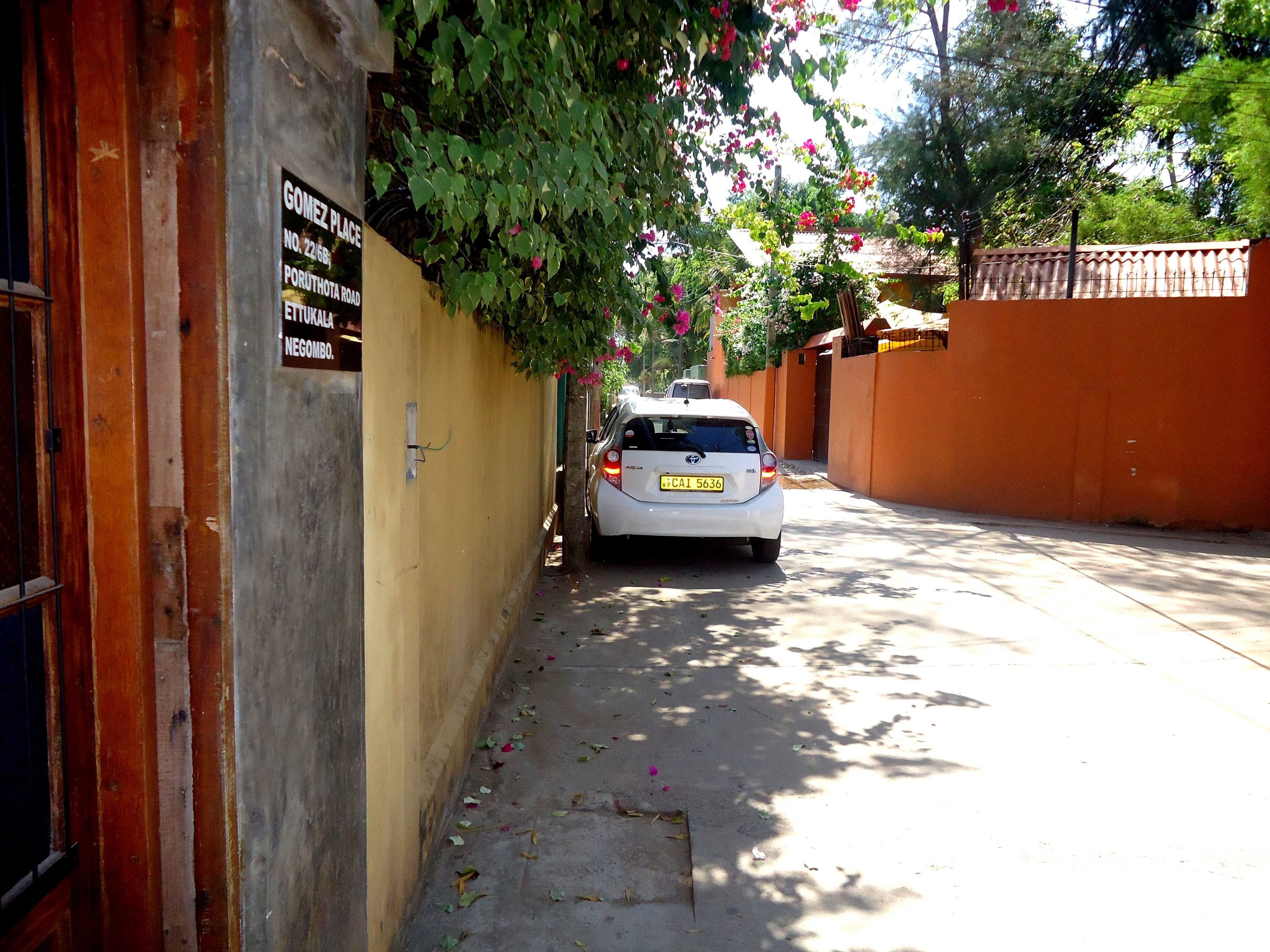 Una calle estrecha con un coche blanco estacionado en la acera, un cartel en la pared izquierda y un edificio con pared roja y una puerta.