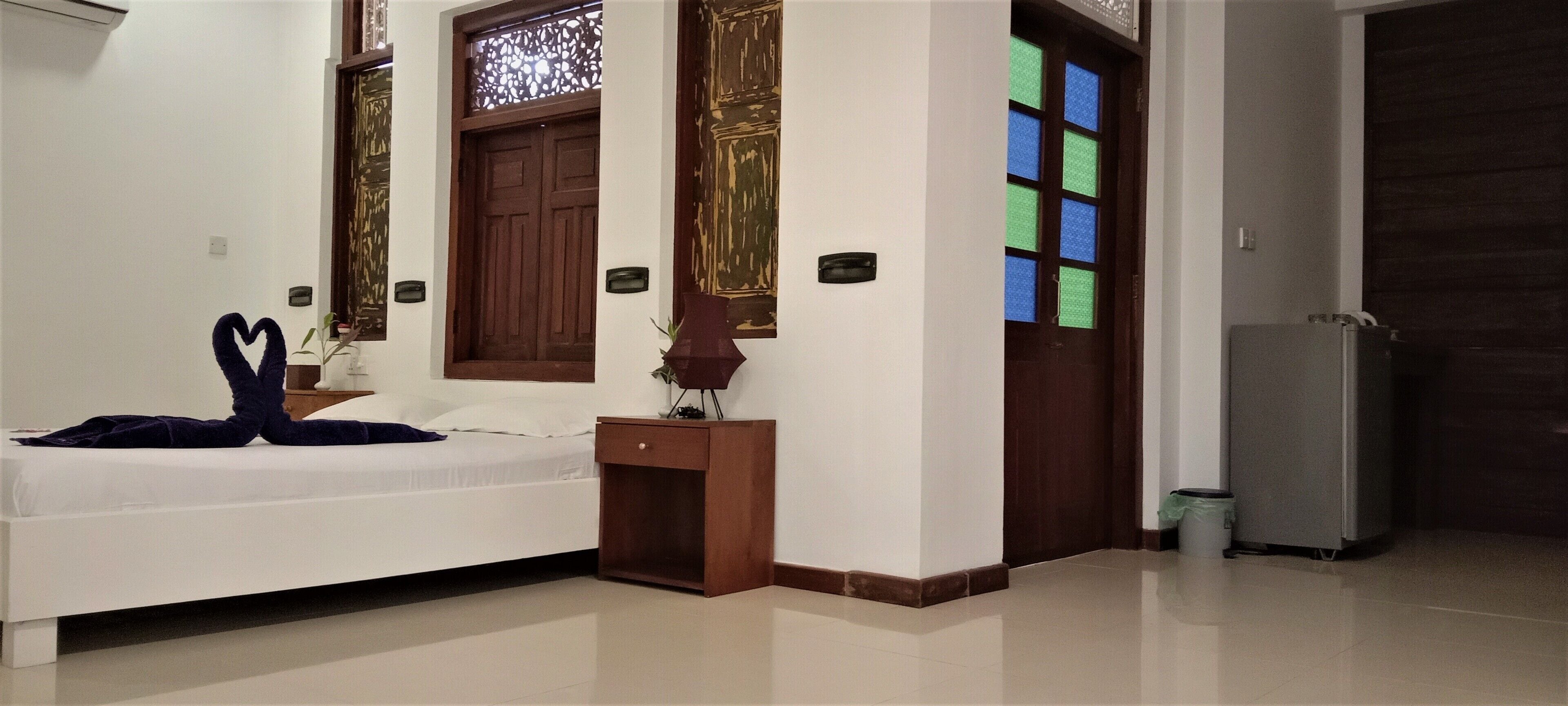 A bedroom with a bed, a nightstand, a dresser, a door with a window, and a refrigerator.