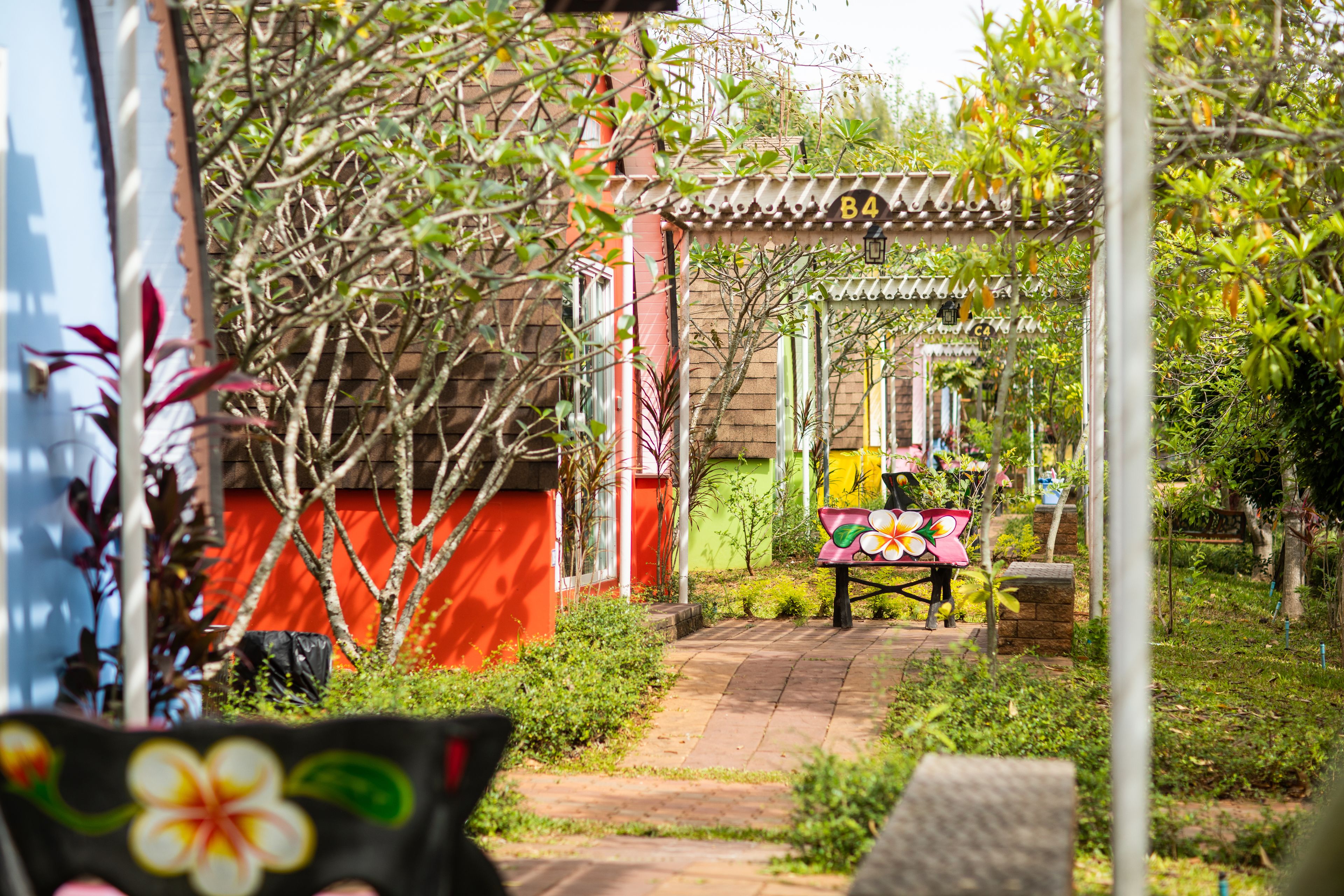 Un sendero de jardín que conduce a un edificio con fachada roja y verde, un área techada con un letrero con el número 'B4', y una colorida exhibición de flores.