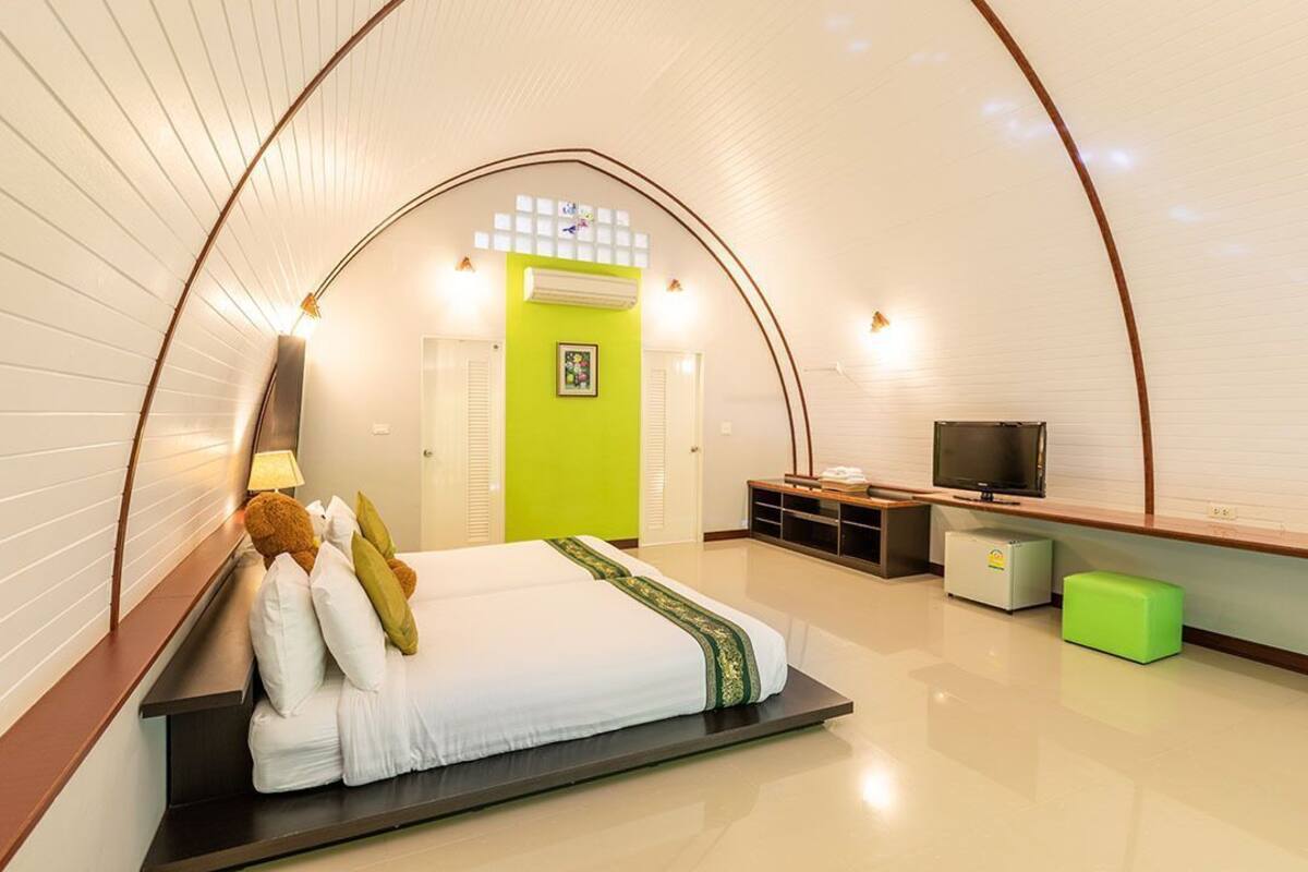 Una habitación de hotel moderna con techo curvo, una cama con almohadas, un televisor, un refrigerador pequeño y un reposapiés verde.