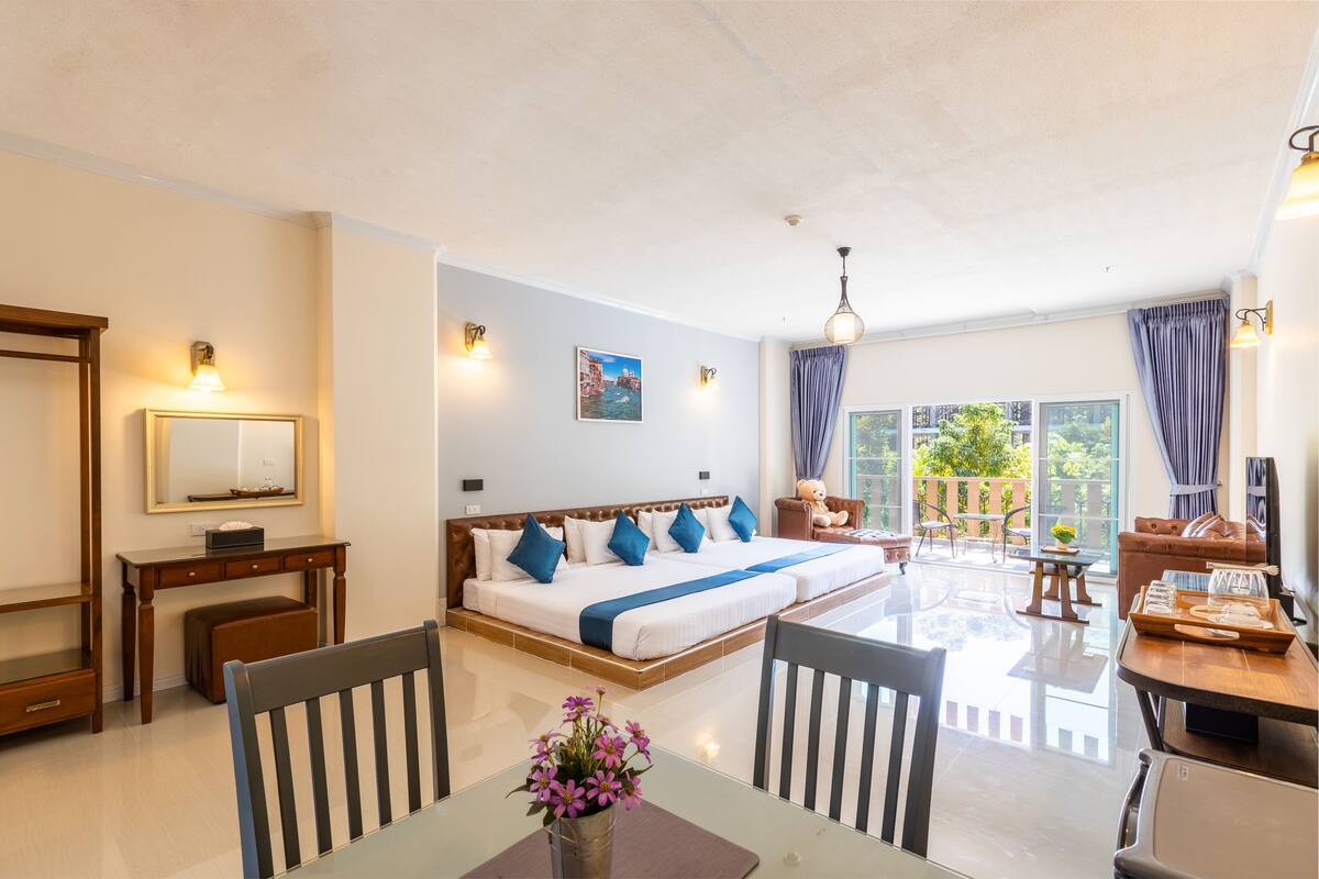 Una habitación de hotel con una cama grande, una mesa de comedor, sillas, una mesita con un jarrón de flores y un ventanal con cortinas.