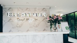 Een receptiebalie met een marmeren ontwerp, erboven de naam "BLUE SKY VILLA" verlicht. Er staat een bloemenvaas en een incheckbord met uitchecktijden en betaalmogelijkheden.