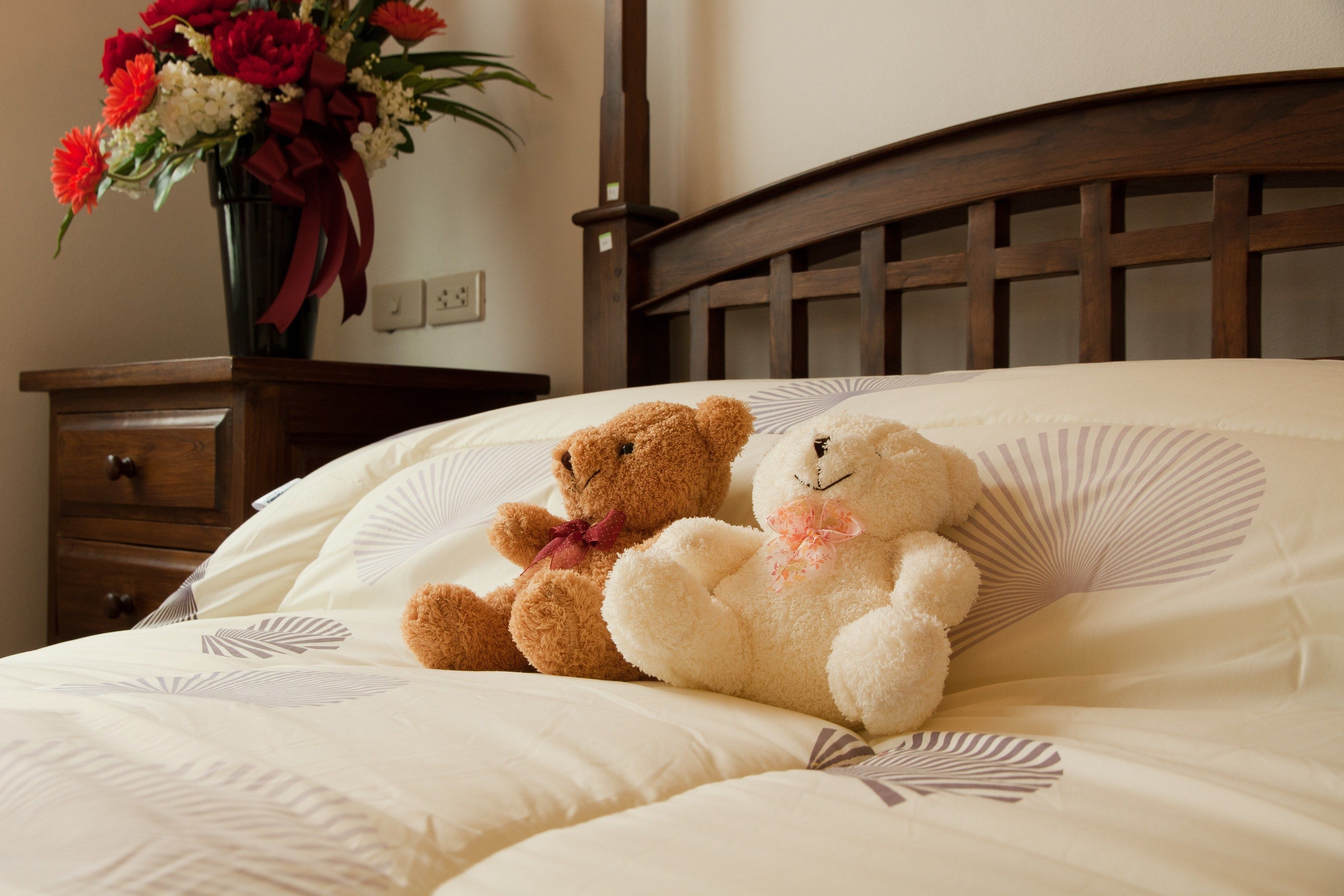 A bed with a wooden headboard, a teddy bear, and a vase of flowers on a nightstand.