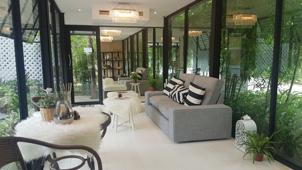 Een moderne woonkamer met een grijze bank, een witte, pluizige poef en rieten stoelen. Er zijn grote ramen met beplanting buiten, en een kroonluchter hangt aan het plafond.