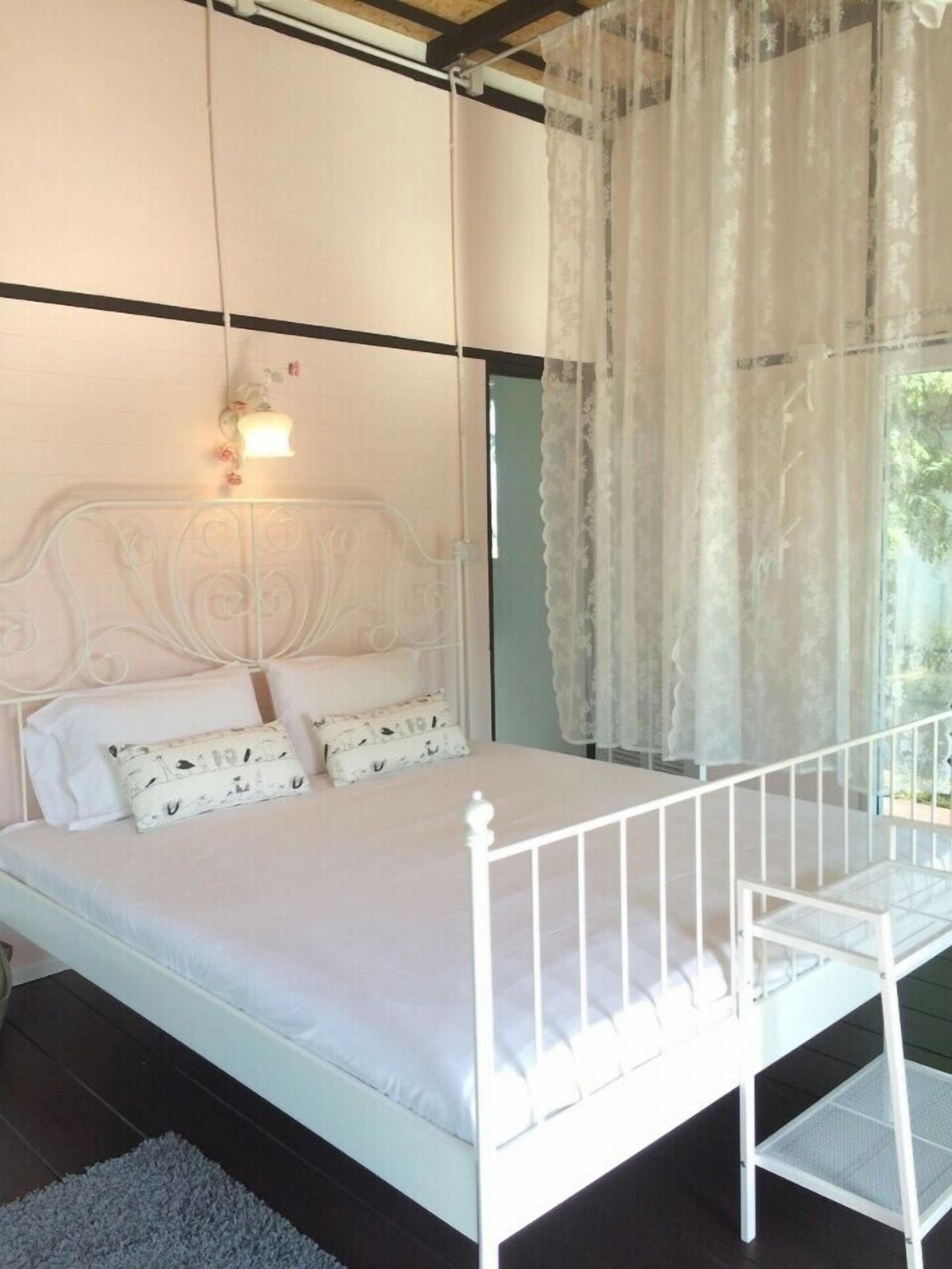 Ein Schlafzimmer mit einem weißen Metallbettgestell, einer weißen Matratze und weißer Bettwäsche. Ein Fenster mit transparenten Vorhängen bietet Blick auf Grünflächen.
