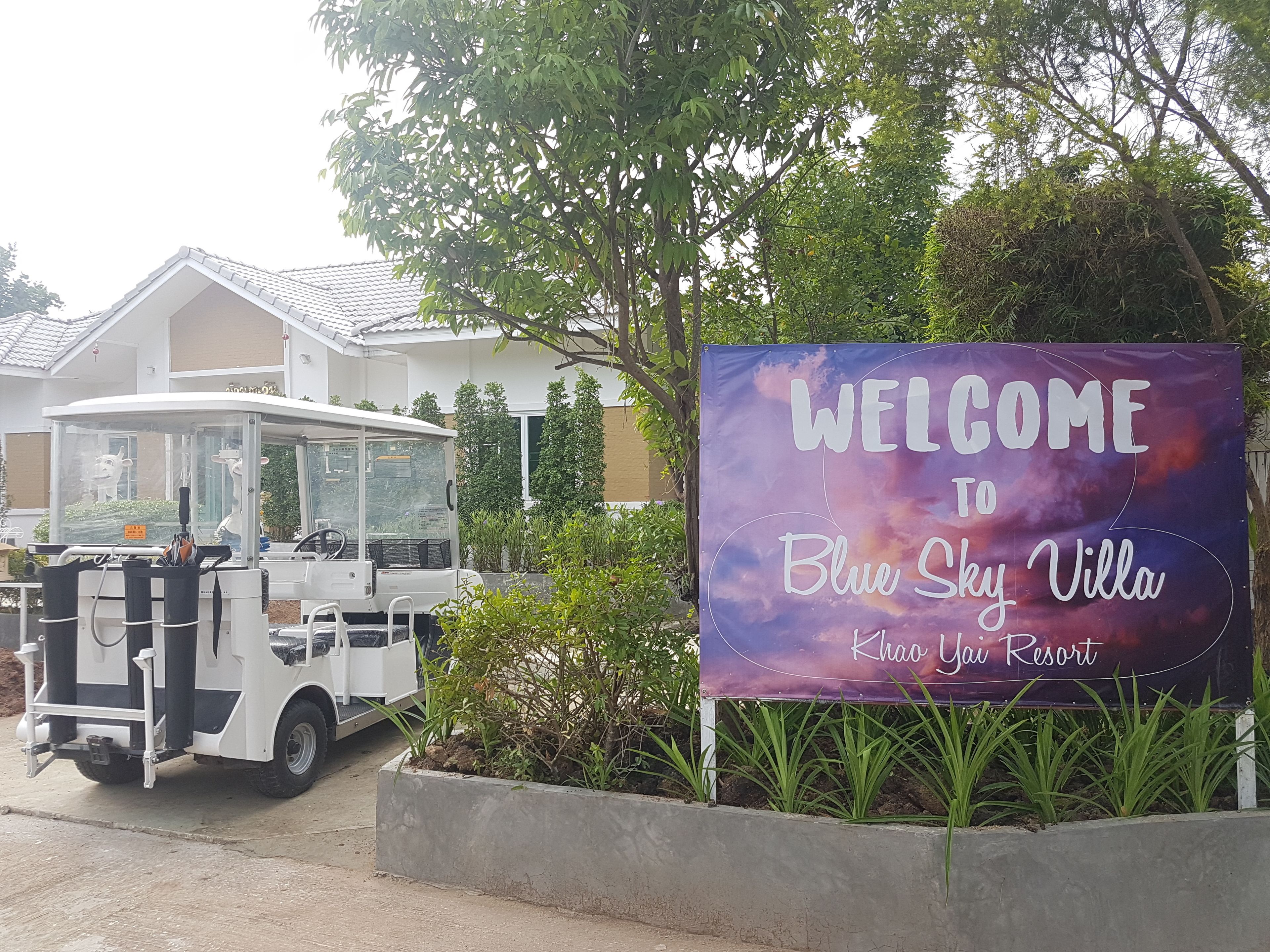 A welcome sign for Blue Sky Villa at Khao Yai Resort, a white vehicle parked nearby, and a building with a tiled roof in the background.