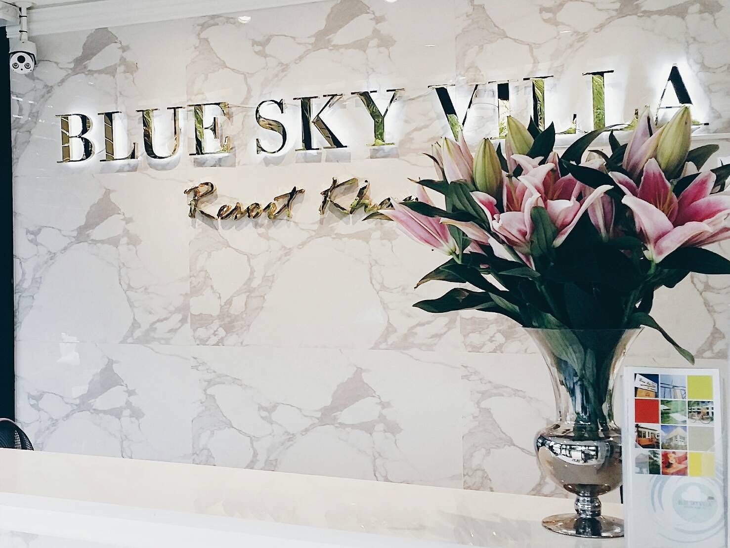 A reception desk with a marble wall background, a sign reading "BLUE SKY VILLA," and a vase with pink flowers.