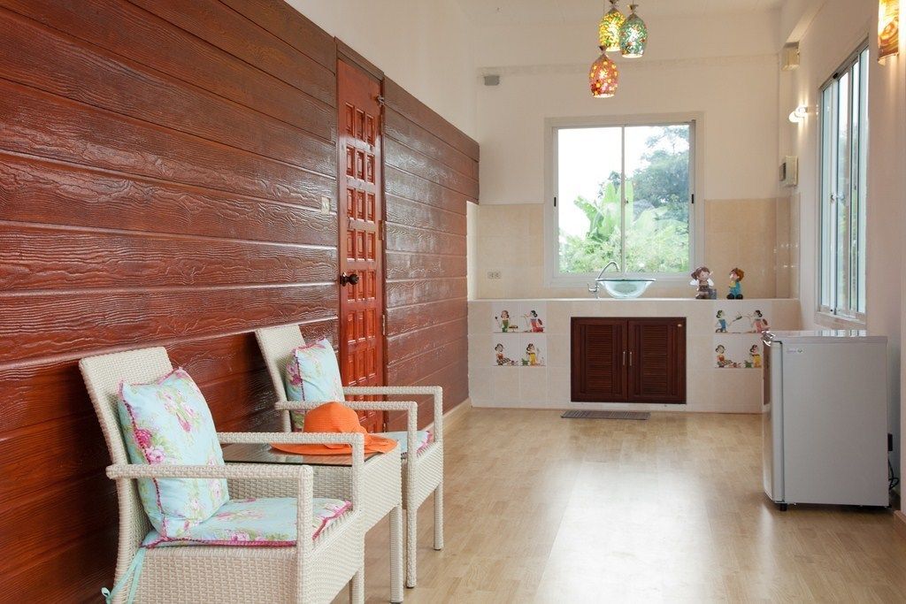 A room with a wooden wall, a white wicker sofa with cushions, a white refrigerator, and a window with a view of greenery.