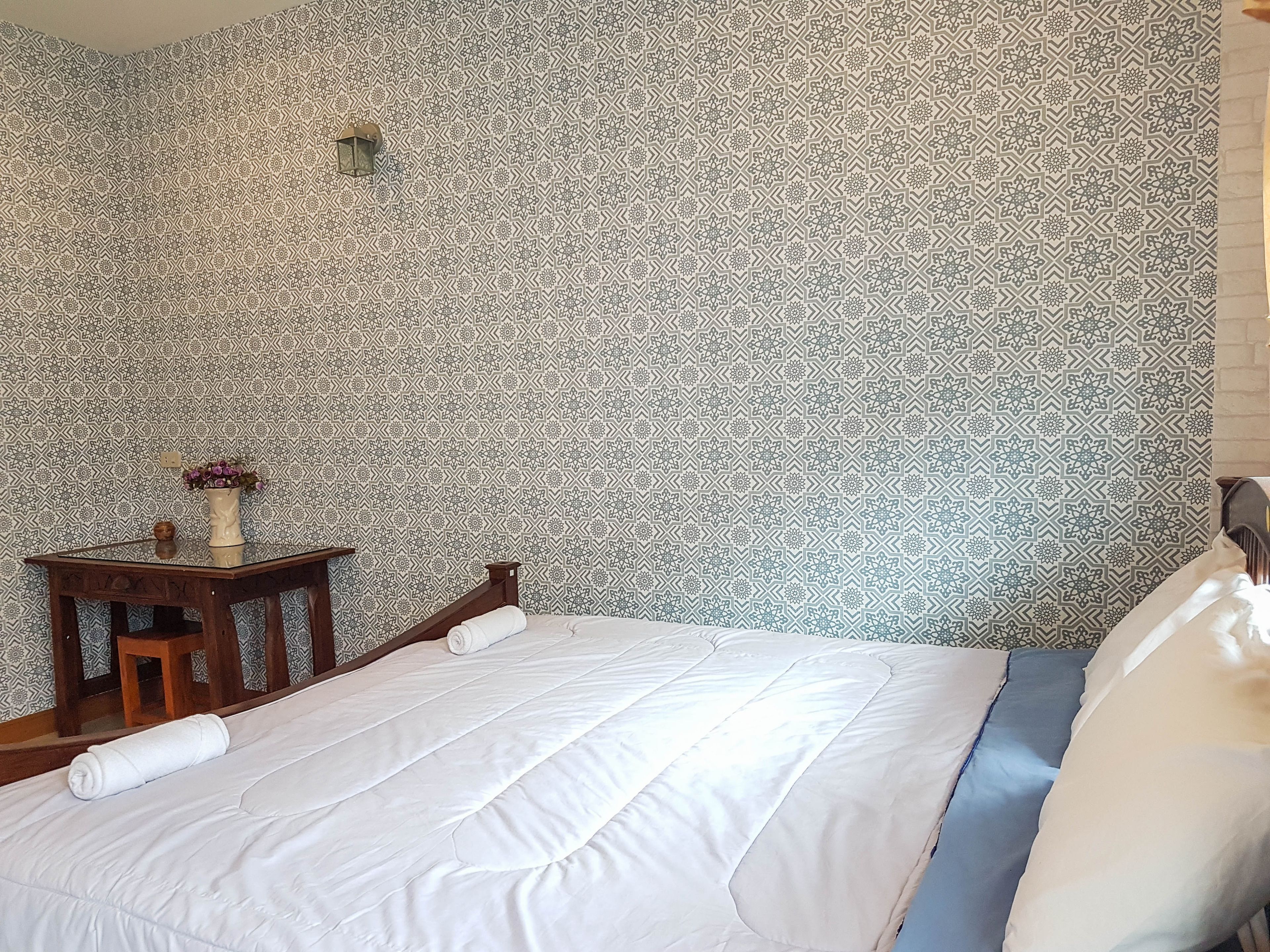 A neatly made bed with white bedding, a small wooden nightstand with a vase of flowers, and a patterned wallpaper.