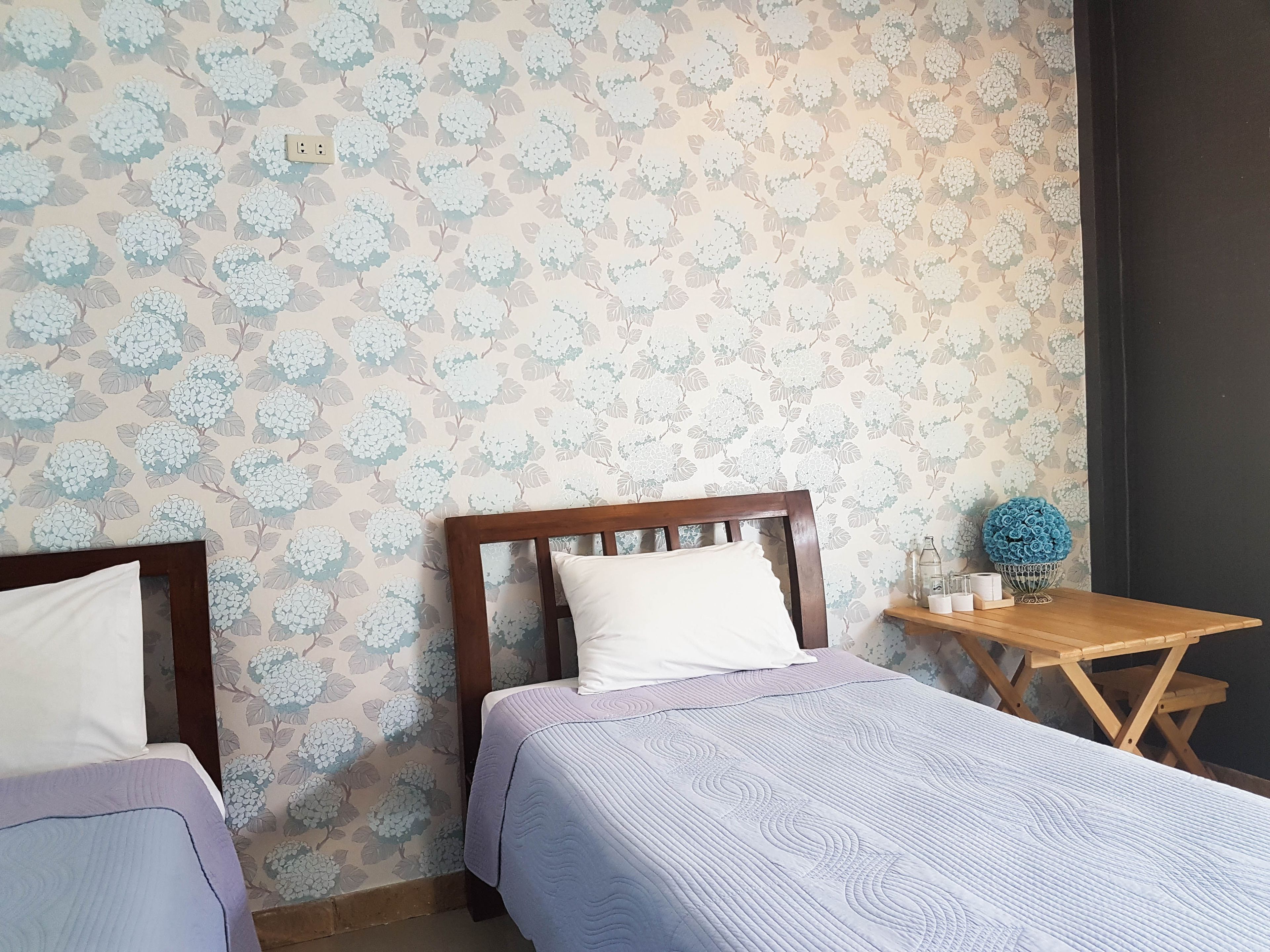A bedroom with two beds, a wooden headboard, a small wooden table with a vase, and a wall with a floral pattern.