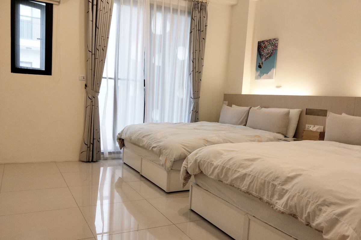 A hotel room with two beds, a large window with sheer curtains, a painting on the wall, and a tiled floor.