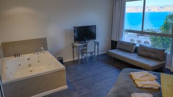 A room with a large bathtub, a desk with a TV, a sofa with cushions, and a view of the sea.