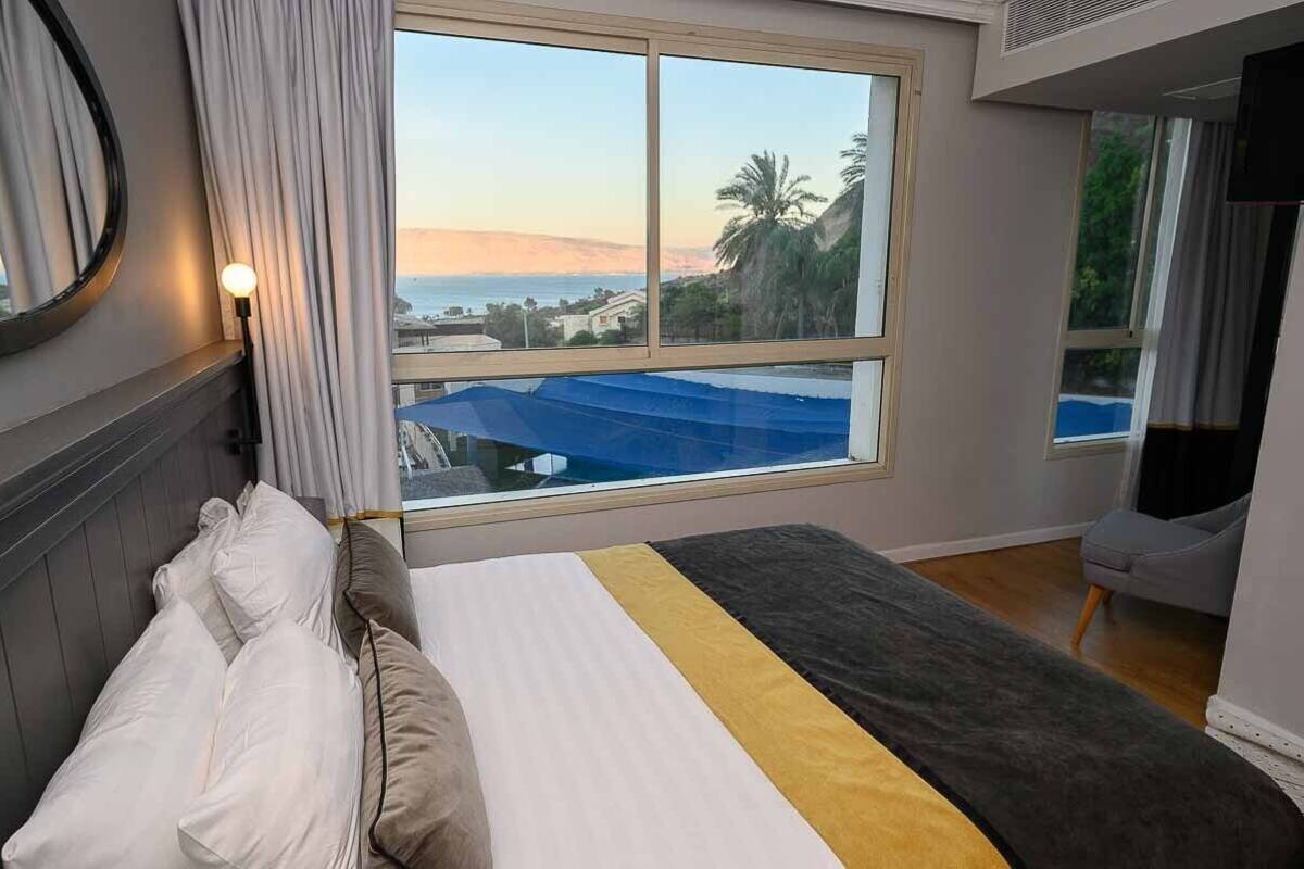 A hotel room with a bed, a large window with a view of the sea and hills, a chair, and a flat-screen TV.