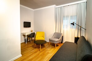 Ein Wohnzimmer mit grauem Sofa, geläufigem Sessel, grauem Sessel, einem kleinen runden Tisch, einer schwarzen Stehlampe, einem weißen Schreibtisch und einem Fernseher an der Wand.