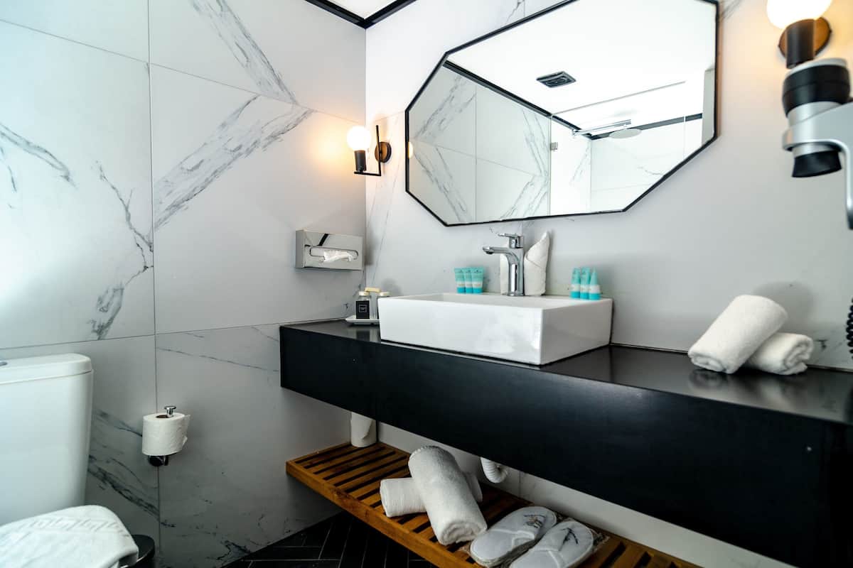 A modern bathroom with a white sink, black countertop, and a large mirror. There are towels on a wooden shelf, a toilet paper holder, and a towel rack.