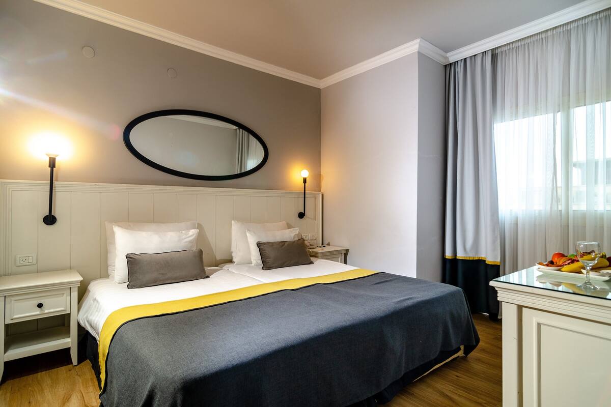 A hotel room with a large bed, two bedside tables, a mirror, and a dining area with a table set for two.