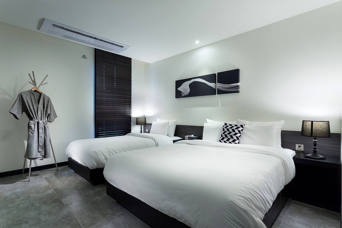 A modern hotel room with two beds, a robe rack, a wall-mounted air conditioner, and a nightstand with a lamp.