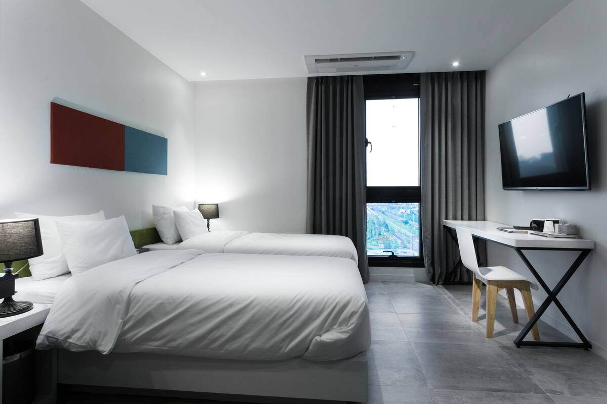 A modern hotel room with a bed, desk, chair, TV, and a view of a cityscape.