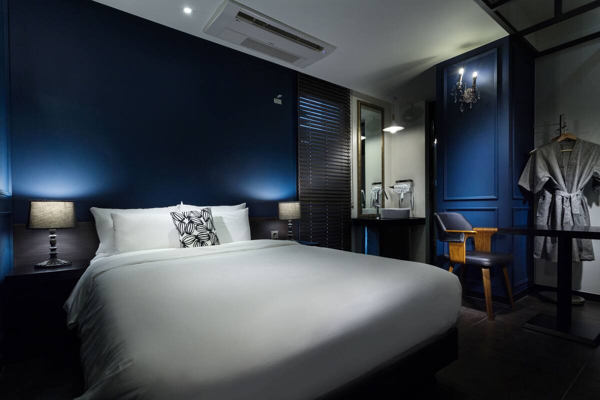 A modern hotel room with a large bed, a desk with a chair, a mirror, and a robe hanging on a rack.