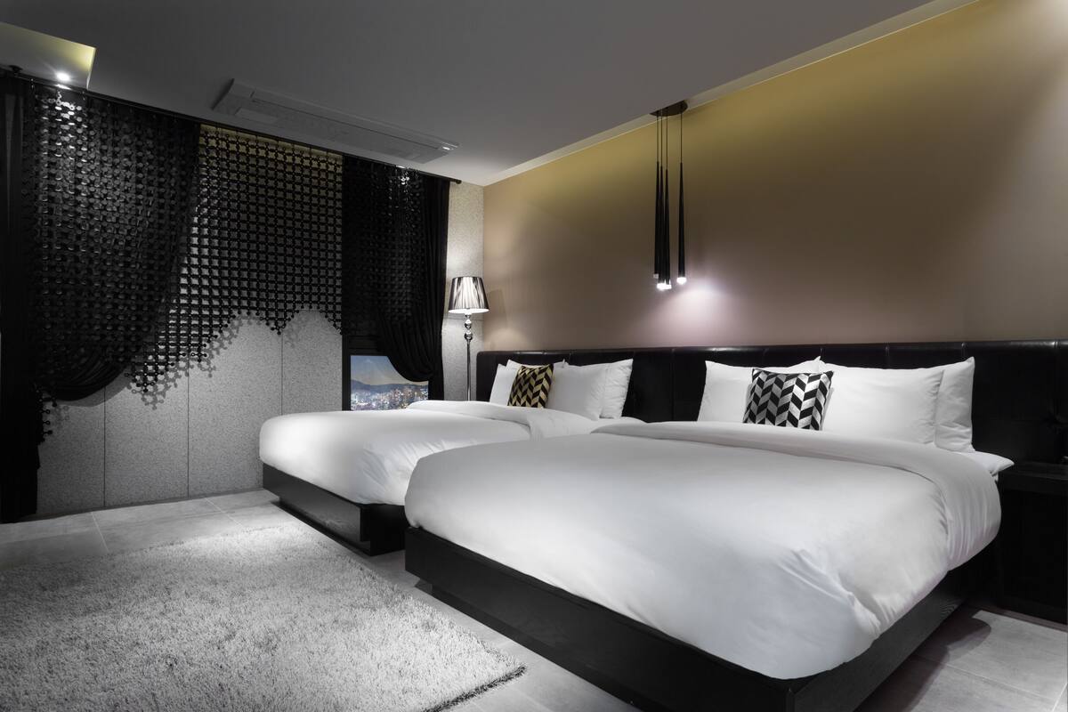 A modern hotel room with two beds, a large window with curtains, a nightstand, and a lamp.