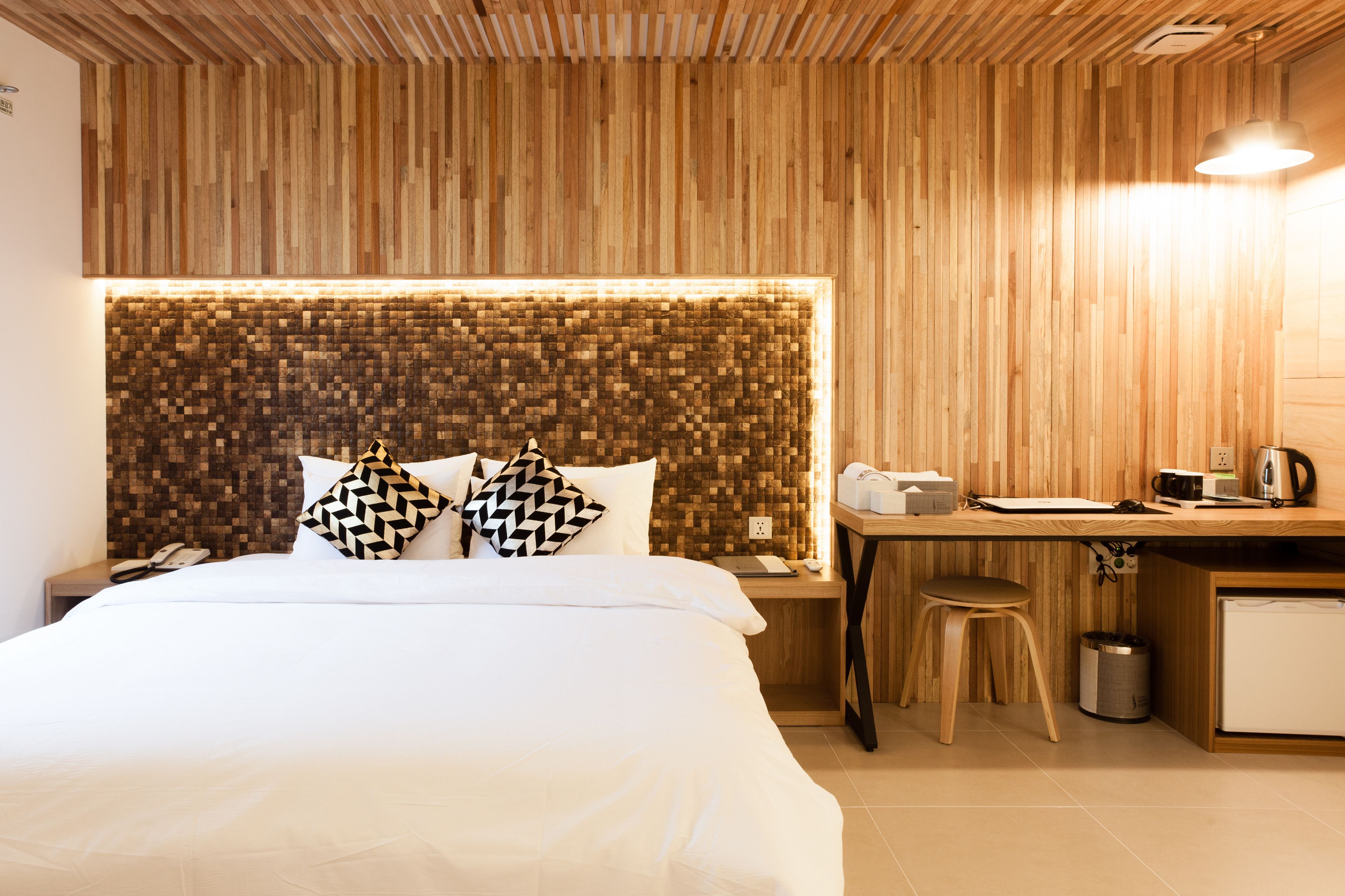 A hotel room with a wooden headboard, a bed with white linens, a desk with a chair, a small kitchenette, and a trash can.